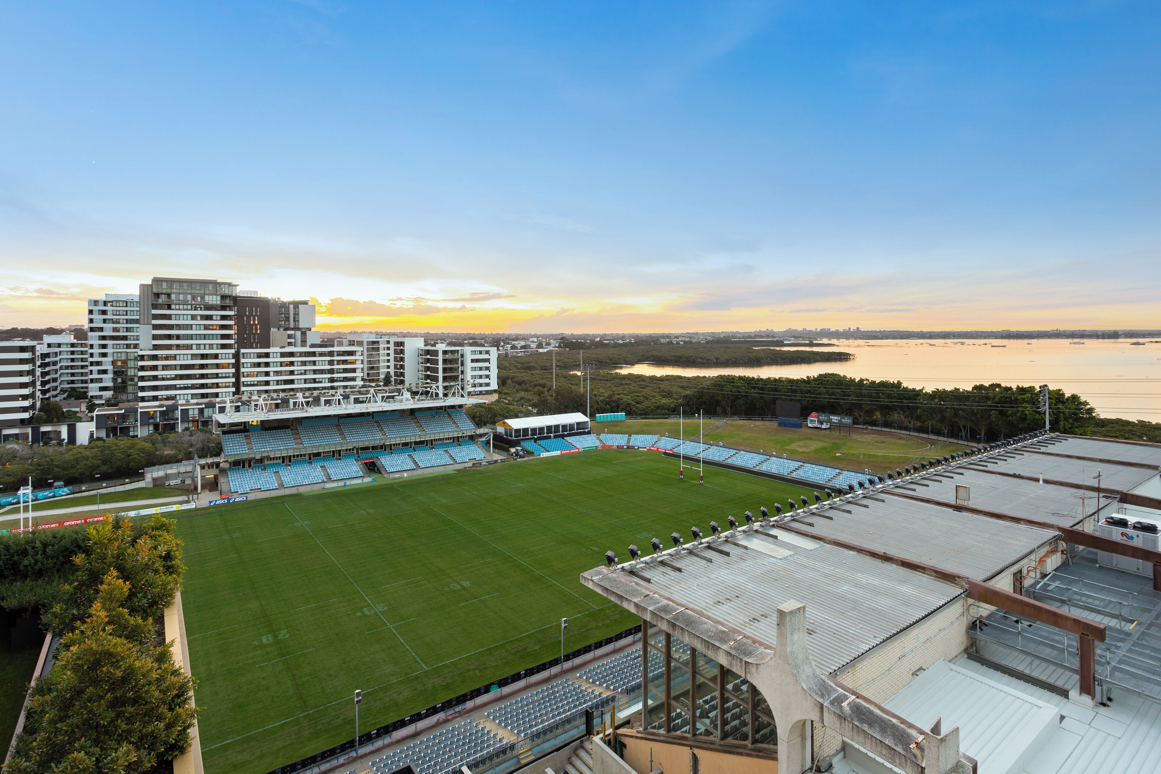 Un estadio con césped verde y asientos azules, rodeado de edificios y una masa de agua.