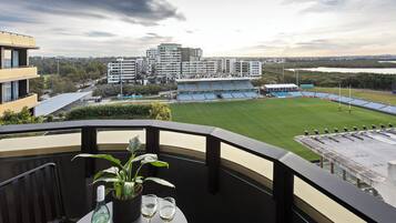 Un balcon avec une table, un pot de fleurs et des verres à vin. Au fond, on aperçoit un stade avec des sièges bleus et un ensemble de bâtiments.