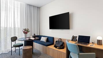 A modern hotel room with a sofa, a desk with a laptop, a chair, a small table with a vase of flowers, and a large window with curtains.