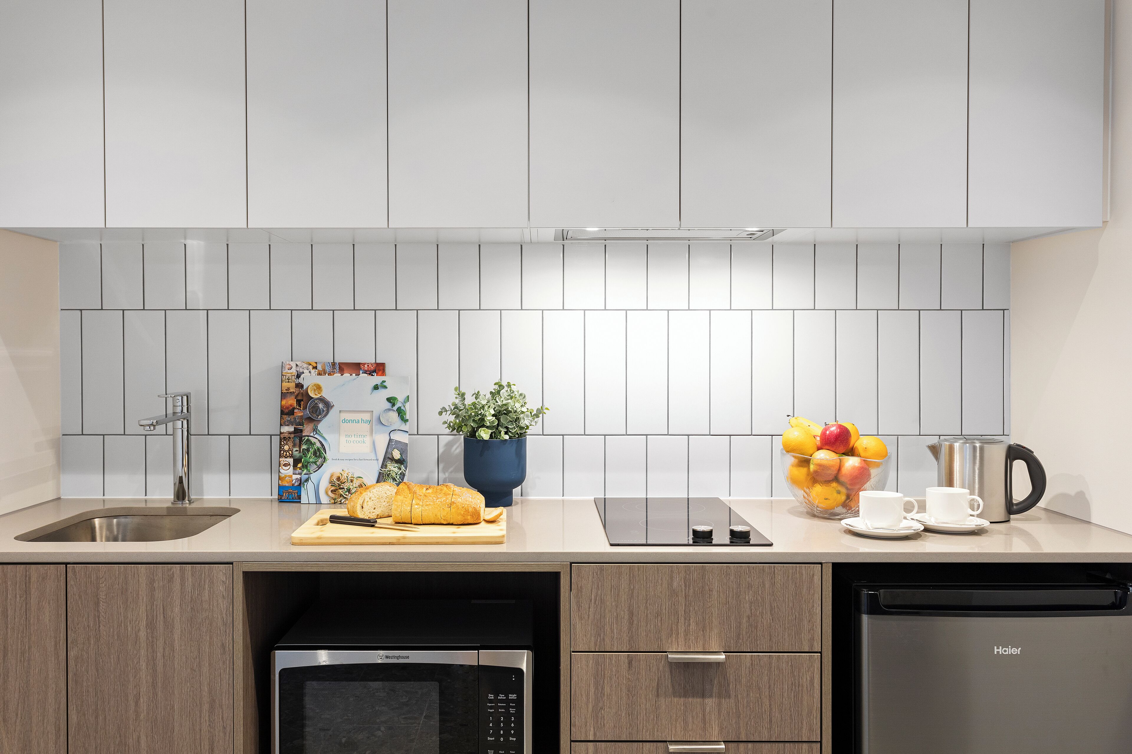 A modern kitchen with a stainless steel sink, a microwave, a dishwasher, and a countertop with a cutting board, a loaf of bread, a potted plant, a bowl of fruit, and a coffee maker.