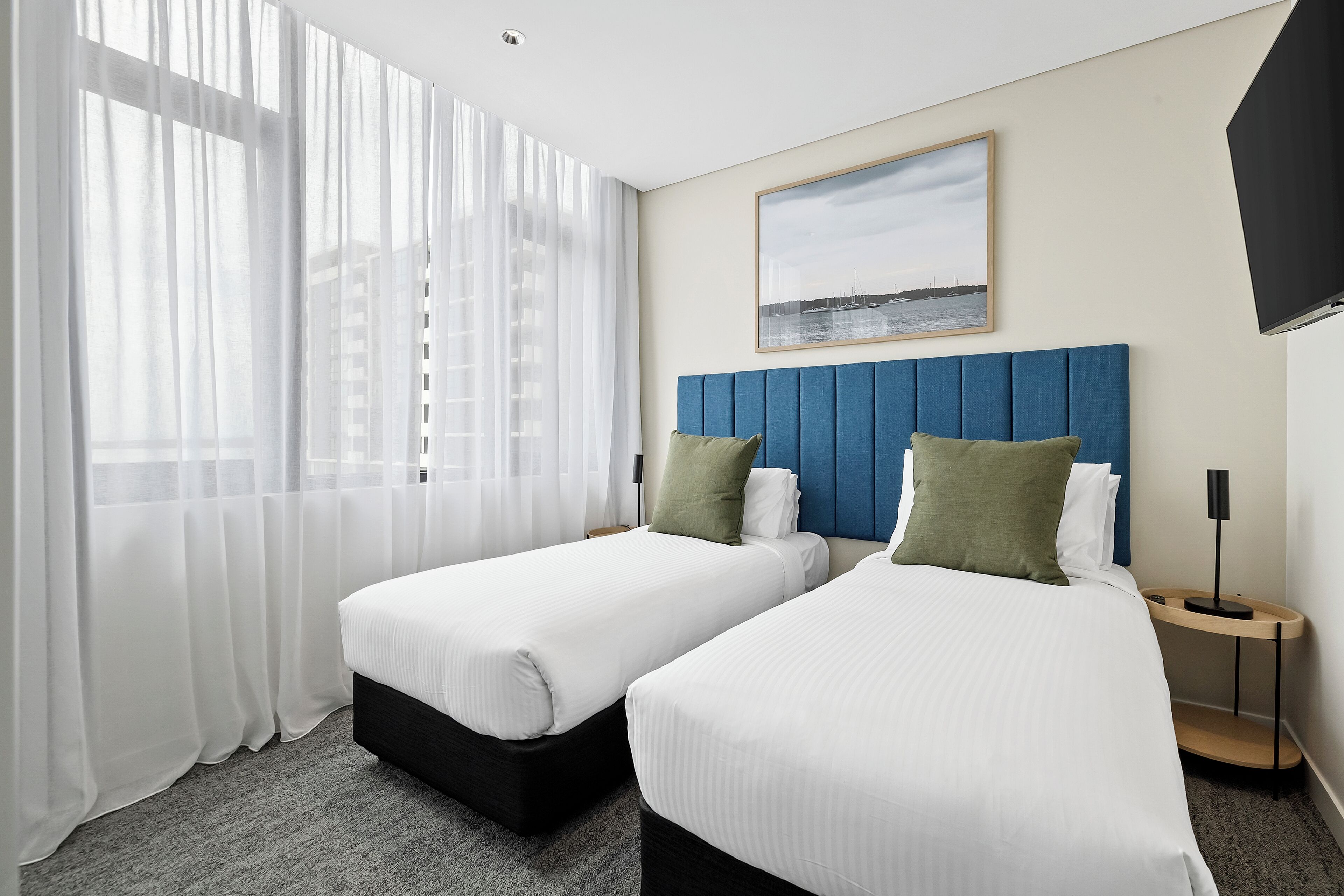 Ein Hotelzimmer mit zwei Betten, einem blauen Kopfteil, weißer Bettwäsche, einem gerahmten Bild an der Wand und einem Nachttisch mit einer Lampe.