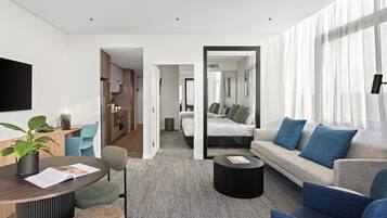 Et moderne hotelværelse med gråt tæppe, en sofa med blå puder, et rundt sofabord, et spisebord med en plante og et tv monteret på væggen.
