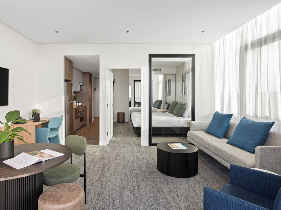 Et moderne hotelværelse med gråt tæppe, en sofa med blå puder, et rundt sofabord, et spisebord med en plante og et tv monteret på væggen.