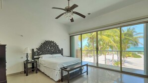 Ein Schlafzimmer mit einem Bett, einem Nachttisch, einer Lampe, einem Deckenventilator und einem großen Fenster mit Blick auf Palmen und den Strand.
