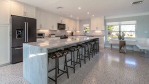 A modern kitchen with white cabinets, stainless steel appliances, a marble island with bar stools, and a large window offering a view of the outdoors. - Waterfront oasis, sunsets, heated pool, hot tub, boat dock, near boat ramp (Bradenton)