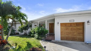 A two-story house with a covered porch, a wooden garage door, and a well-maintained lawn with various plants. - Jungle house Heated Salt pool, 5 min away from the beach! (Boynton Beach)