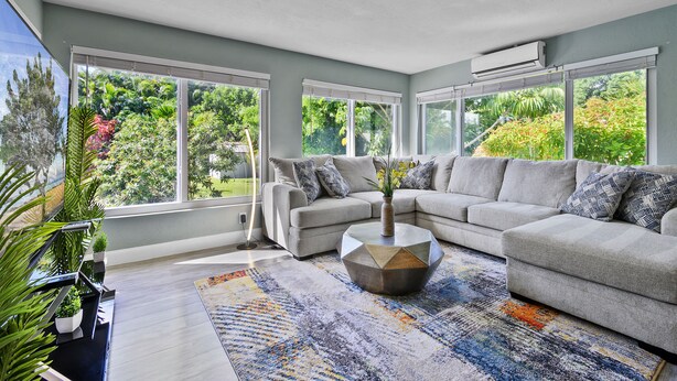 A modern living room with a grey sofa, a coffee table, and a large window offering a view of greenery. - Jungle house Heated Salt pool, 5 min away from the beach! (Boynton Beach)