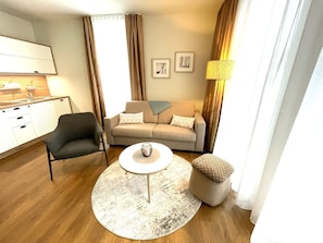 Een moderne woonkamer met een bank, fauteuil, salontafel, poef en een kleed. Er is een keuken met een spoelbak en kastjes, en een lamp op een bijzettafeltje.