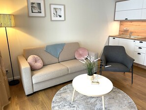 Un salon comprenant un canapé, un fauteuil, une table basse, ainsi qu’un coin cuisine avec des placards et un évier.