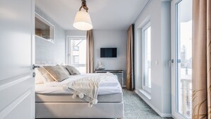 A modern hotel room with a large bed, a flat-screen TV, a desk with a lamp, and a view of the city through large windows. - Haus Zingst, Appartement 24 für bis zu 4 Personen. - Haus Zingst (Fuhlendorf)