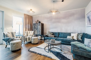 Un salon moderne comprenant un canapé, des fauteuils, un pouf et une table basse. On y trouve également un meuble avec un motif en chevrons et une lampe.