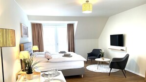 A modern hotel room with a bed, bedside table, lamp, chair, TV, and a small dining area. - Haus Zingst, Appartement 31 für bis zu 2 Personen. - Haus Zingst (Fuhlendorf)