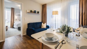 Une chambre d’hôtel moderne dotée d’un coin repas, d’un canapé bleu, d’une table à manger dressée avec des assiettes, des verres et des couverts, et offrant une vue sur l’extérieur grâce à de grandes fenêtres.