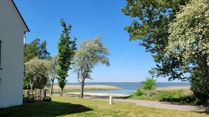 A grassy area with a pathway leading to a body of water, surrounded by trees and a white building. - Haus Barth, Appartement 12 für bis zu 4 Personen. - Haus Barth (Fuhlendorf)