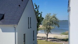 A white building with a sloped roof, a tree in the foreground, and a body of water in the background. - Haus Barth, Appartement 19 für bis zu 4 Personen. - Haus Barth (Fuhlendorf)