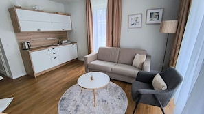 Ein modernes Wohnzimmer mit einer Couch, einem runden Couchtisch und einem Sessel. Es gibt eine Küchenzeile mit Schränken und einem Wasserkocher.