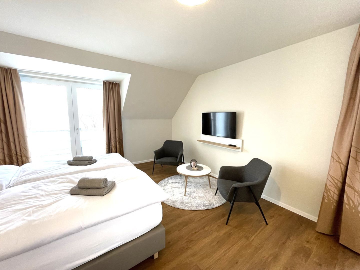 A modern hotel room with a large bed, a TV mounted on the wall, two armchairs, and a small round table.