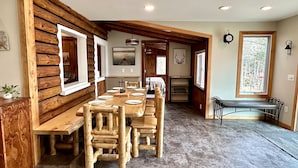 A log cabin interior with a long dining table, wooden benches, a bed, and a window with a view of trees.