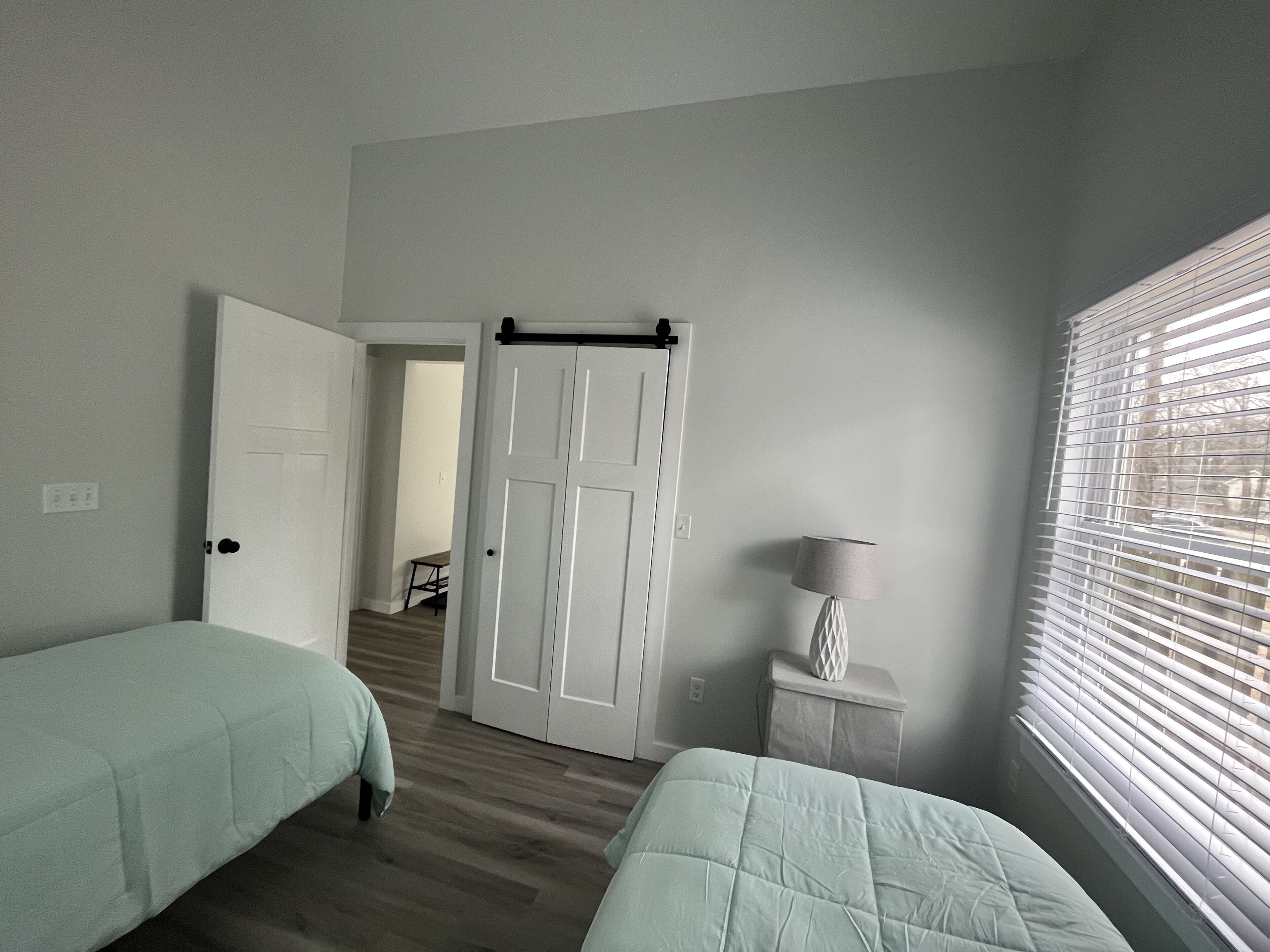 Une chambre avec deux lits, une porte donnant sur une autre pièce, une table de chevet avec une lampe et une fenêtre avec des stores.