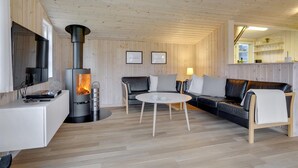 A cozy living room with a wood-burning stove, a sofa, a coffee table, and a kitchen area with shelves. - Charming holiday home close to water (Sæby)