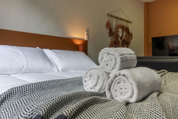 A neatly made bed with white linens and rolled towels, a patterned throw blanket, and a wall-mounted decor piece.