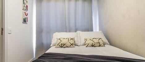 A bedroom with a bed, two pillows, a blanket, and a curtain.