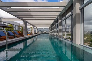 Une piscine intérieure moderne avec des parois en verre transparent, des chaises longues et une vue sur la ville.