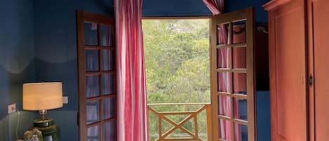 A bedroom with a large bed, a nightstand with a lamp, a wooden wardrobe, and a view of a wooden deck and greenery through open French doors.