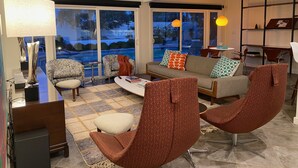 Una sala de estar moderna con sofá, sillones, una mesa de centro y televisor. Amplias ventanas ofrecen vistas a una piscina y árboles.