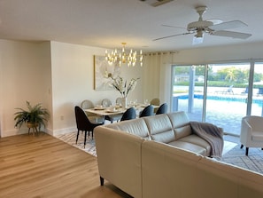 Una sala de estar moderna con un sofá color beige, una mesa de comedor y sillas. Se ve una ventana grande con vistas a una piscina.
