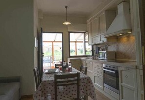 A kitchen with white cabinets, a dining table with a polka dot tablecloth, and a view of the outside through large windows. - Appartamento a 200 metri dal mare Granchio (Alghero)