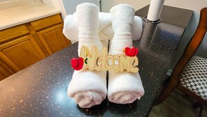 A pair of white towels with a wooden sign reading "Welcome" and two red flowers, placed on a speckled countertop in a room with wooden cabinets and a chair with a patterned cushion. - Renovated Suite with 1 queen bed and a Twin. Kitchen & bath for your privacy. (Goldsboro)