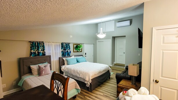 A bedroom with two beds, a wooden floor, a chandelier, a TV, and a door. - Renovated Suite with 1 queen bed and a Twin. Kitchen & bath for your privacy. (Goldsboro)