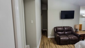 A living room with a leather recliner, a flat-screen TV mounted on the wall, and a wooden floor. - Renovated Suite with 1 queen bed and a Twin. Kitchen & bath for your privacy. (Goldsboro)