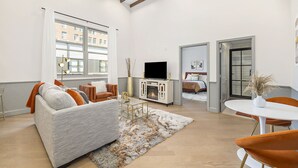 A modern living room with a sofa, coffee table, and TV. There is a fireplace, a dining area with a table and chairs, and a large window with curtains. - Iconic Morris Ave condo retreat sleeps 6 +rooftop (Birmingham)