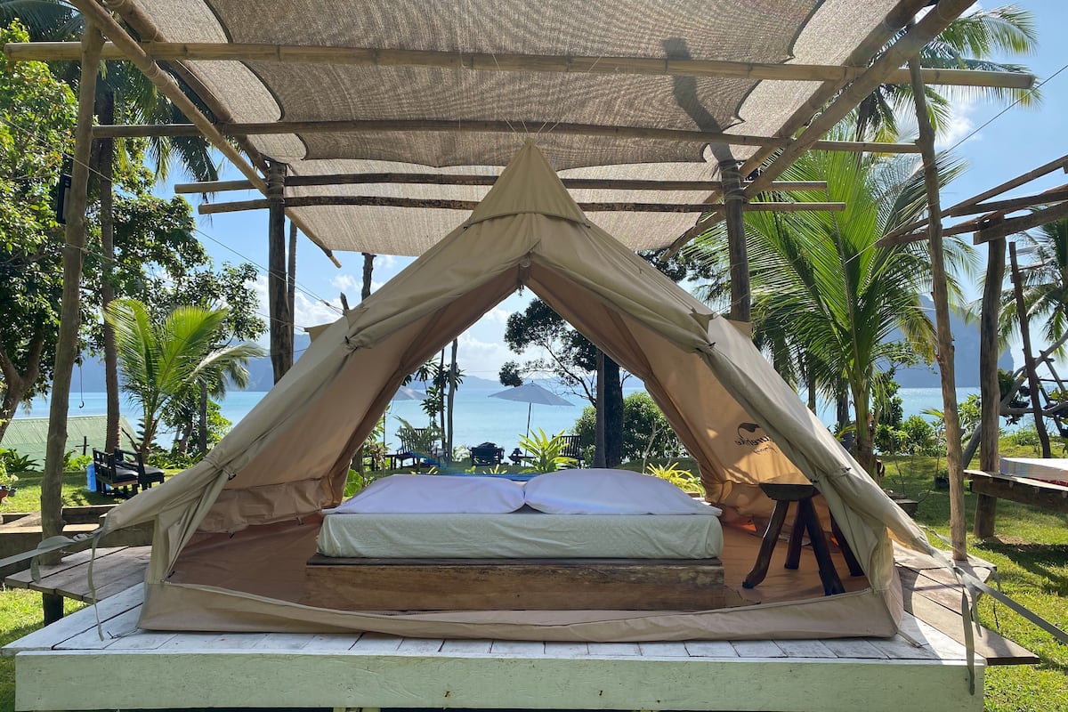 A tented accommodation with a bed, surrounded by palm trees and a view of the sea.