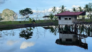 Una casa sobre pilotes sobre el agua con reflejo nítido, rodeada de palmeras y una montaña al fondo.