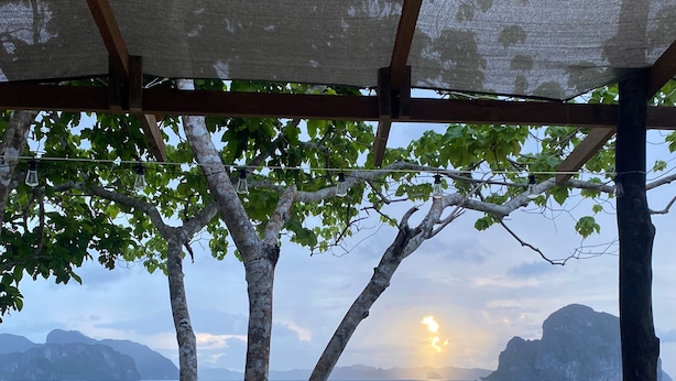 Terraza de madera con vistas al mar y montañas, una lámpara colgante y plantas en macetas.