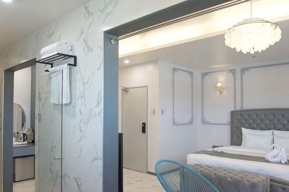 A modern hotel room with a glass shower enclosure, a bed with a tufted headboard, and a chandelier.