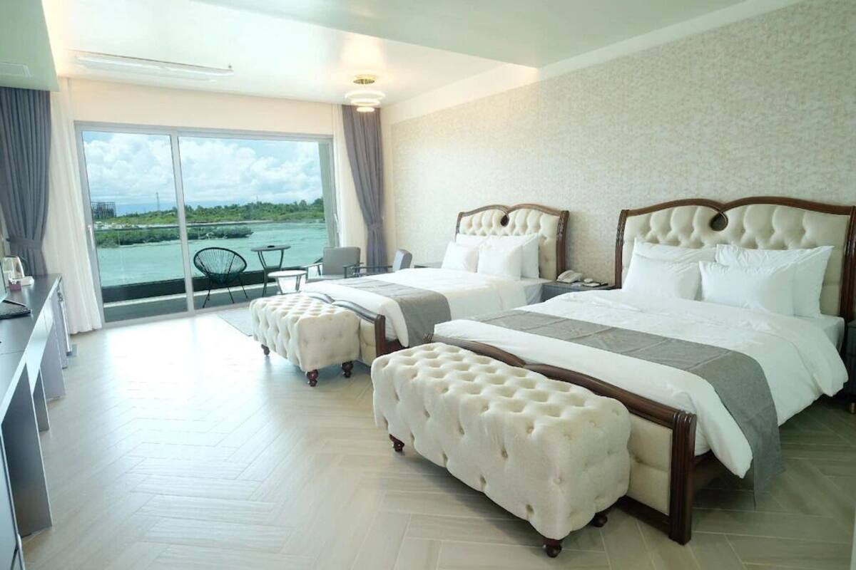 A hotel room with two beds, a balcony with a view of the water, and a desk with a chair.