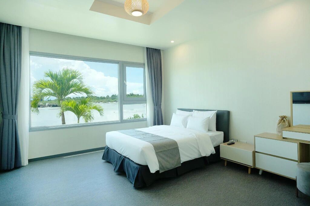 Een moderne hotelkamer met een groot bed, een nachtkastje, een kaptafel en uitzicht op palmbomen door een groot raam.