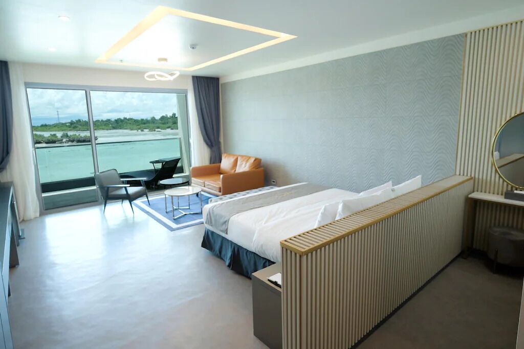 A modern hotel room with a large bed, a seating area with a chair and table, a view of the sea through large windows, and a balcony.