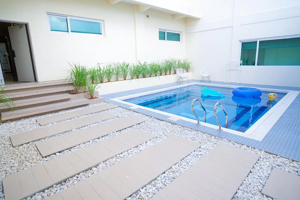 Piscina con escalones de acceso, rodeada por un edificio blanco con ventanas y un pequeño jardín.