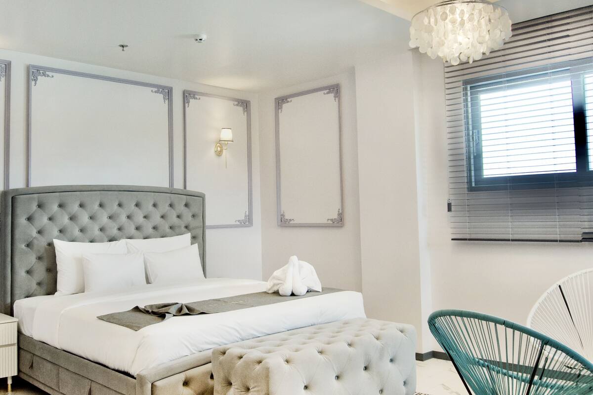A modern bedroom with a tufted headboard, a large bed, a bedside table, a chair, and a window with blinds.