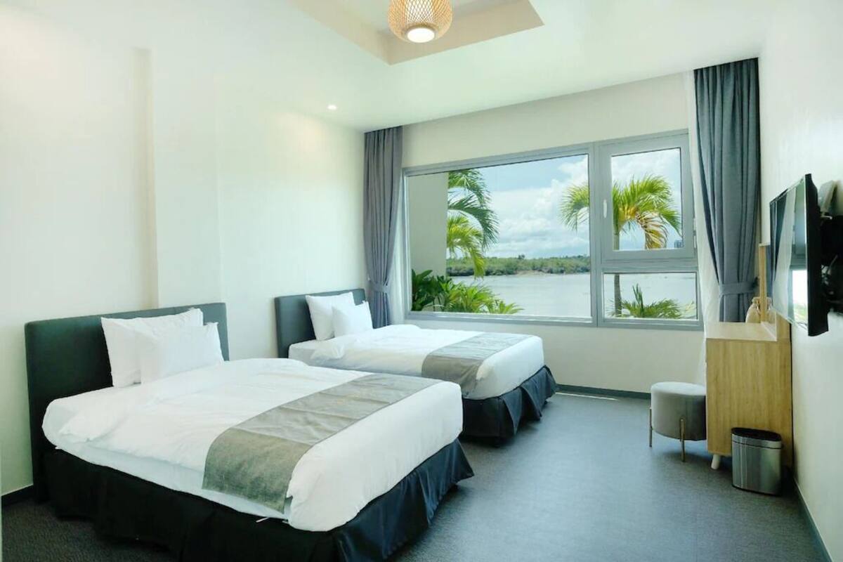 A hotel room with two beds, a desk, a TV, and a window with a view of palm trees and a body of water.