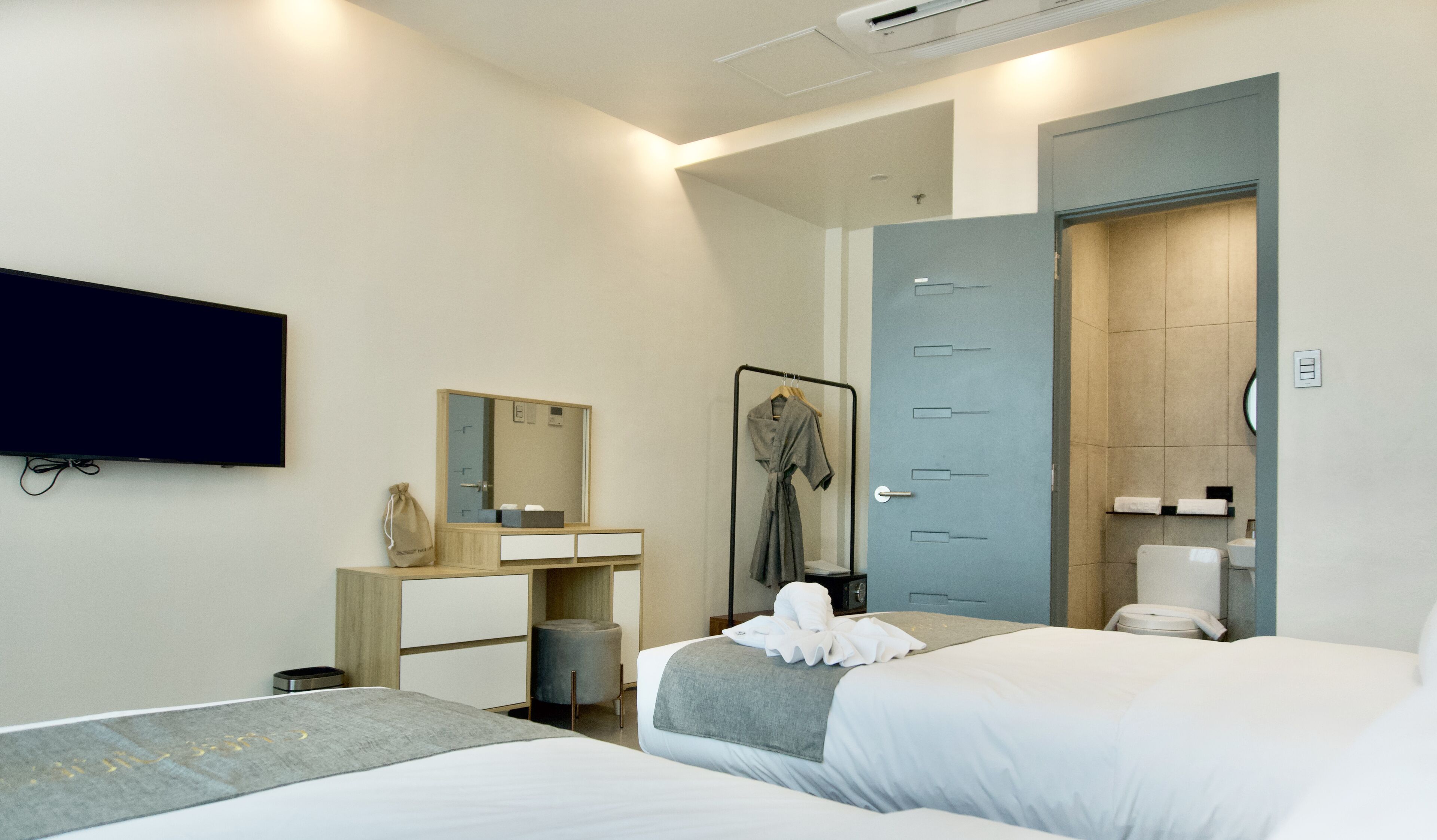 A hotel room with two beds, a desk, a mirror, a robe, and a bathroom with a toilet and sink.