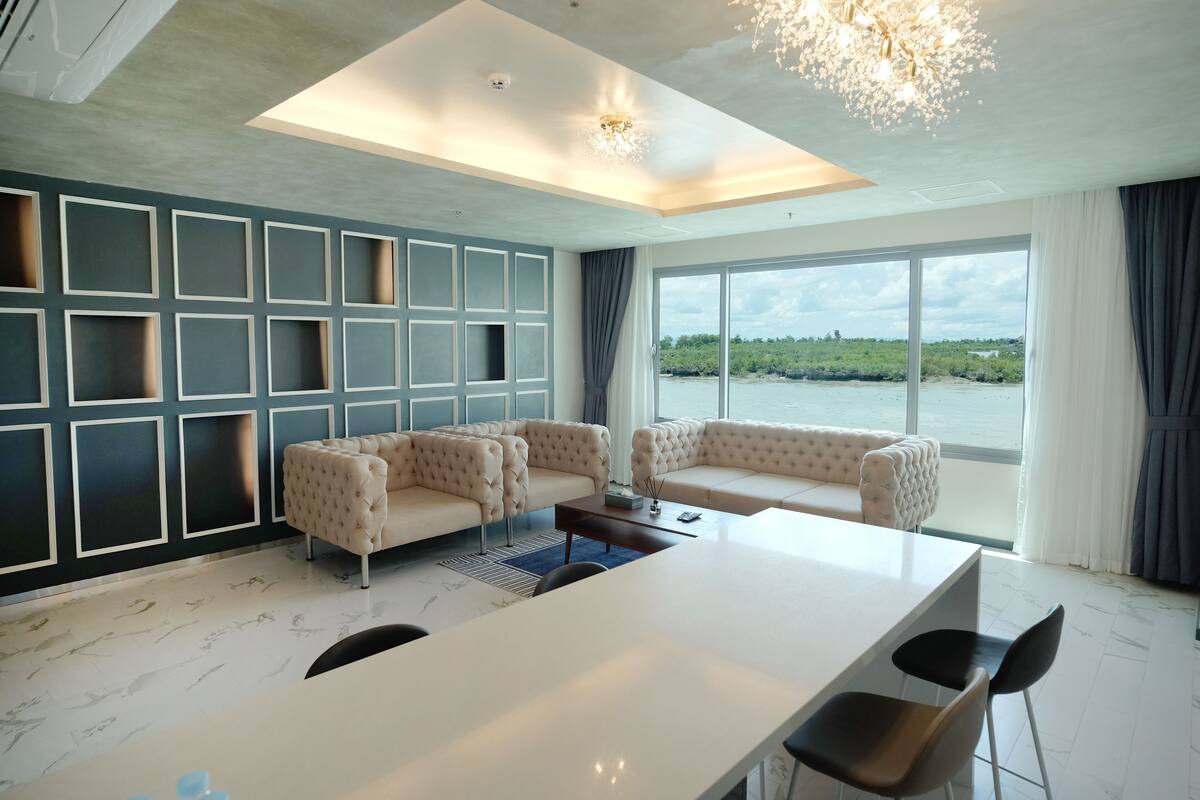 A modern living room with a large white table, beige sofas, and a view of a body of water through large windows.