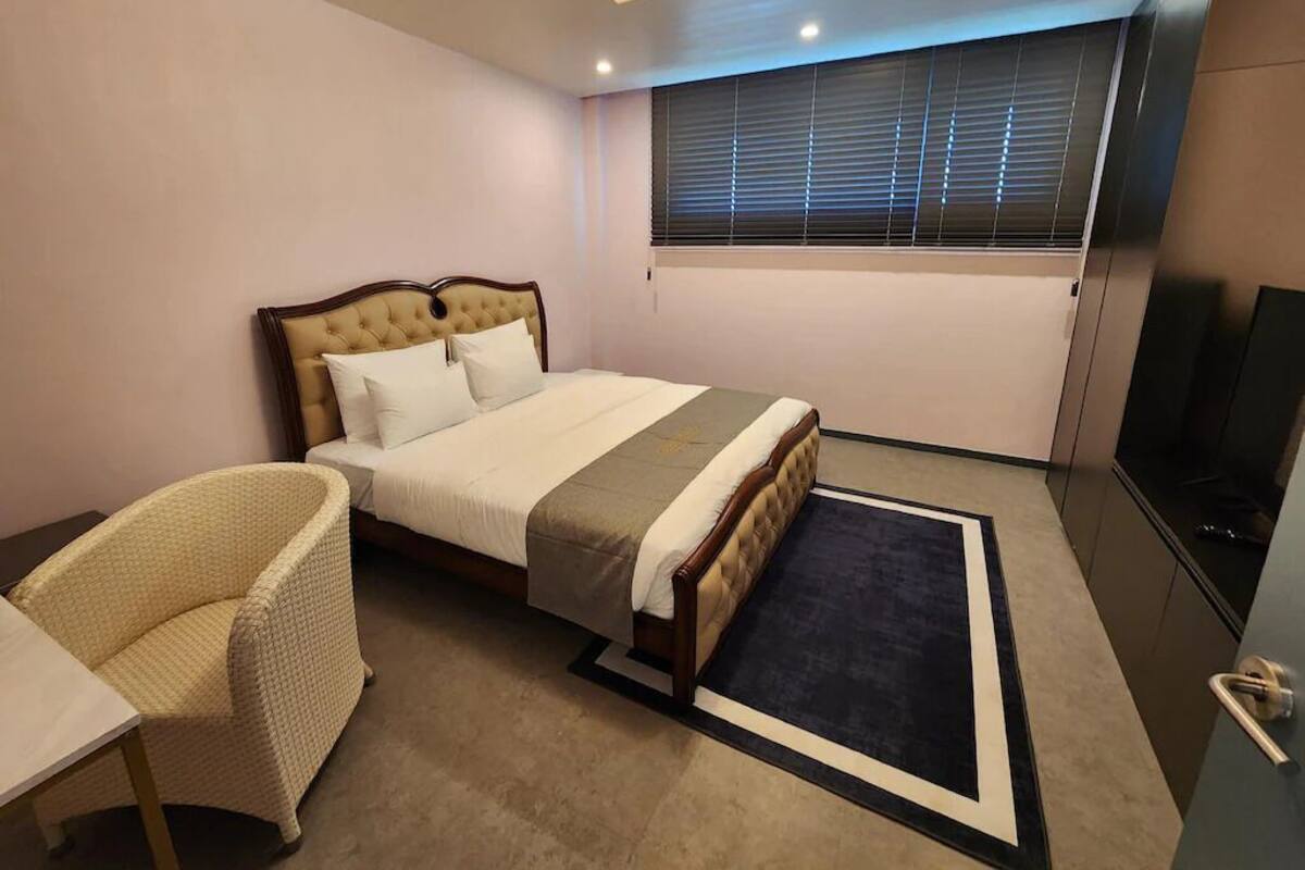 A bedroom with a bed, a chair, a TV, and a window with blinds.