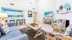 Un moderno salón con una mesa de centro de vidrio, un sofá y un sillón de colores. Se ve una cocina al fondo con armarios blancos y una pared de acento azul.