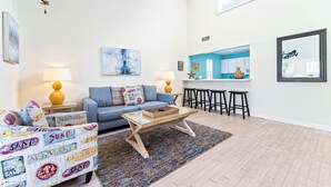 Una sala de estar con un sofá azul, una mesa de centro de vidrio y un sillón estampado. Hay una zona de cocina con encimera azul y taburetes.